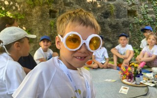 Ein Junge mit orangen Brillengläsern lächelt während er mit anderen Kindern auf einer Wiese sitzt. Im Hintergrund befinden sich Blumen und Spielzeug.
