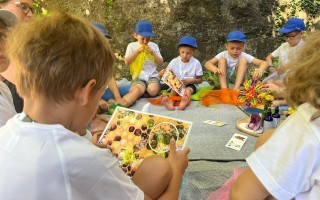 Kinder sitzen auf einer Decke und schauen gemeinsam ein Bild von Früchten an. Es ist ein sonniger Tag.