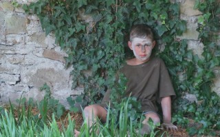 Ein Junge mit blasser Gesichtsfarbe sitzt inmitten von Gräsern vor einem bröckelnden Mauerwerk.