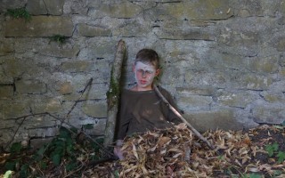 Junge hält Stock vor bröckelnden Mauer. Laub bedeckt den Boden.