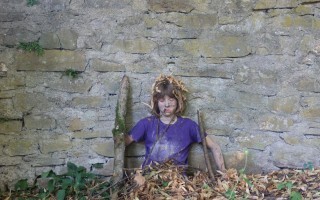 Ein junges Mädchen mit Haarkranz sitzt auf einem Holzpfosten zwischen Laub und einem bröckelnden Steinmauer.