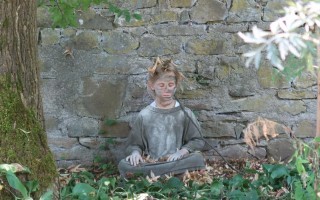 Eine grüne Statue eines Kindes sitzt in einer lotusartige Haltung vor einem Steinmauer. Laub liegt im Vordergrund.