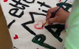 Eine Person malt mit Farbe auf ein rotes Blatt Papier. Es sind stilisierte Formen und Herzen in Schwarz dargestellt.