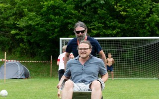 Mann fährt Rollstuhl auf grüner Wiese mit Fußballtor und Zäunen.