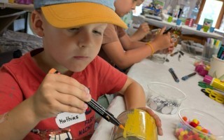 Ein Junge mit Baseballcap malt in einer Glasflasche mit grüner Farbe. Im Hintergrund weitere Kinder beim Basteln.
