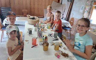 Kinder basteln an einem Tisch mit Farben, Gläsern und Materialien. Ein Tablett mit Papierfetzen steht daneben.