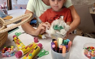 Eine Frau und ihr Sohn sitzen an einem Tisch mit Stiften, Filz und Süßigkeiten. Der Sohn hält eine Schachtel mit Süßigkeiten in der Hand.