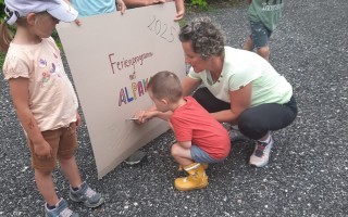 Kinder untersuchen ein Schild mit Buchstaben. Im Hintergrund eine Frau.