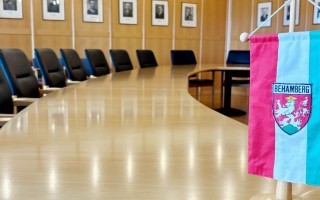 Tischkonferenzraum mit langen, schlichten Tischen und Stühlen. Ein blau-weißes Banner mit Wappen auf dem Tisch.