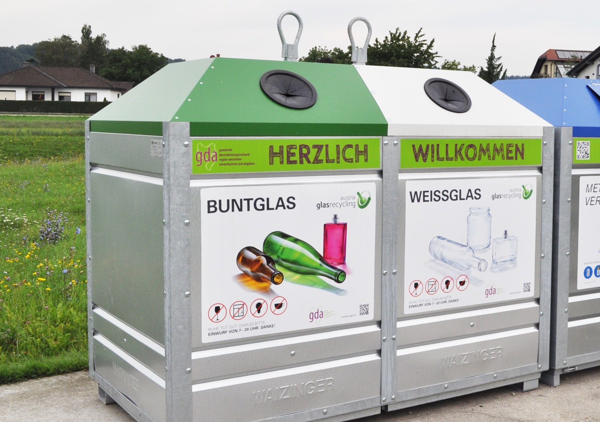 Zwei graue Wertstoffbehälter für buntes und weißes Glas. Auf den Behältern sind entsprechende Logos und Hinweise zur Mülltrennung angebracht.