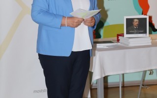 Eine Frau in einem hellblauen Anzug steht vor einem Bücherregal in einer Raum.