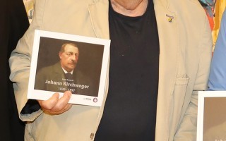 Mann mit grauem Anzug und Gehstock hält ein Porträt von Johann Kirwögerer.