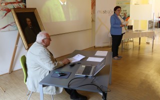 Mann sitzt an einem Schreibtisch mit Laptop und Papieren vor einem abgewandten Stillleben.