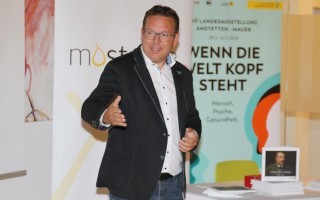 Ein Mann in einem Anzug und Hemd steht vor einem hellen Hintergrund, möglicherweise in einem Büro oder einer Konferenzraum.