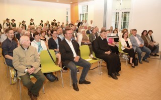 Eine Gruppe von Menschen sitzt in einem Raum auf grünen Stühlen. Einige tragen Mäntel, andere Kleidung verschiedener Art.