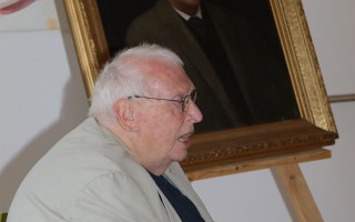 Alter Mann sitzt am Tisch vor einem Holzbild. Er trägt eine Brille und wirkt vertieft in sein Schriftstück.