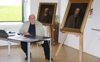 Alter Mann sitzt an einem Tisch mit einem Porträt und einem Laptop. Im Hintergrund steht ein Porträtbild auf einem Podest.