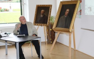 Alter Mann sitzt an einem Tisch in einem Raum. Vor ihm steht ein Bilderrahmen mit einem Porträt.