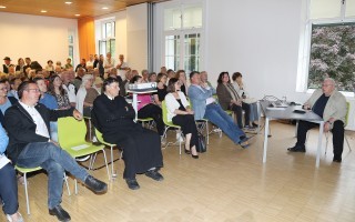 Versammlung von älteren Menschen in einem Raum, sitzend an Tischen, einige mit Getränken und Unterlagen.