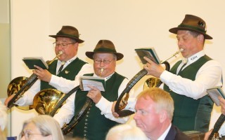 Fünf Männer in Trachten spielen Blechblasinstrumente, darunter ein Trompetenhörner, während sie Notenblätter halten.