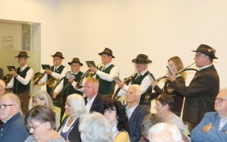 Ein Musikorchester in Trachten spielt vor einer Gruppe älterer Menschen. Die Musiker tragen dunkle Anzüge und Hüte.