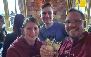 Drei Personen, ein Mann und zwei Frauen, trincken Gläser mit Flüssigkeit. Im Hintergrund sind weitere Personen und eine Holzwand sichtbar.