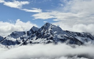 Berglandschaft mit schneebedeckten Gipfeln, umgeben von dichten Wolken am blauen Himmel.