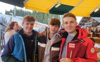 Drei junge Männer sitzen an einem Barhocker in einem Skigebiet. Sie lächeln in die Kamera.