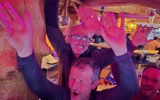 Ein Mann mit grauem Haar und dunkler Jacke lacht mit offenen Händen in einer belebten Bar. Im Hintergrund sind weitere Personen zu sehen.