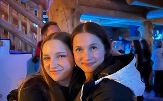 Zwei junge Frauen lachen zusammen in einem belebten Raum. Im Hintergrund verschwommen weitere Personen und Holzbalken.