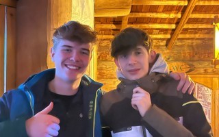 Zwei junge Männer mit blonden Haaren lächeln und zeigen dabei die Daumen nach oben. Sie stehen vor einem Holzwandpaneel.