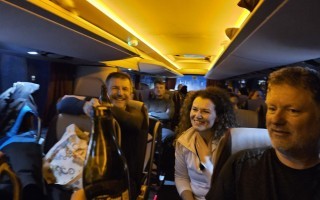 Menschen sitzen in einem Reisebus und lächeln. Im Hintergrund sind weitere Fahrgäste zu sehen.