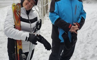 Zwei Skiurlauber in Schneeanzügen mit Skiern vor einem gelben Wegweiser.