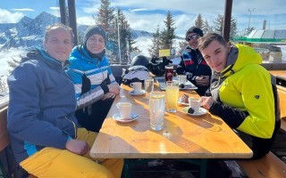 Vier Personen sitzen an einem rustikalen Tisch in einer Skihütte. Im Hintergrund verschneite Berge.