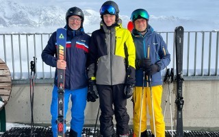 Drei Skifahrer in blauen und gelben Winterjacken mit helblauen Mützen und schwarzen Skibrillen stehen auf einer überdachten Terrasse vor verschneiten Bergen.
