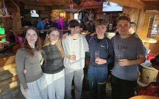 Mehrere junge Männer und eine junge Frau sitzen in einem belebten Bar-Ambiente. Sie halten Gläser in der Hand.