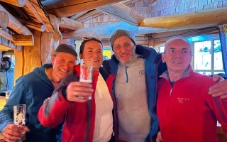 Vier Männer sitzen an einem Tisch und stoßen mit Gläsern an. Sie lächeln und wirken freundlich.