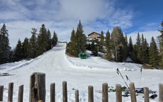 Schneespotten auf Skipiste mit Zauberteppich und Wägelchen, umgeben von Nadelbäumen und einem Alpenchalet.