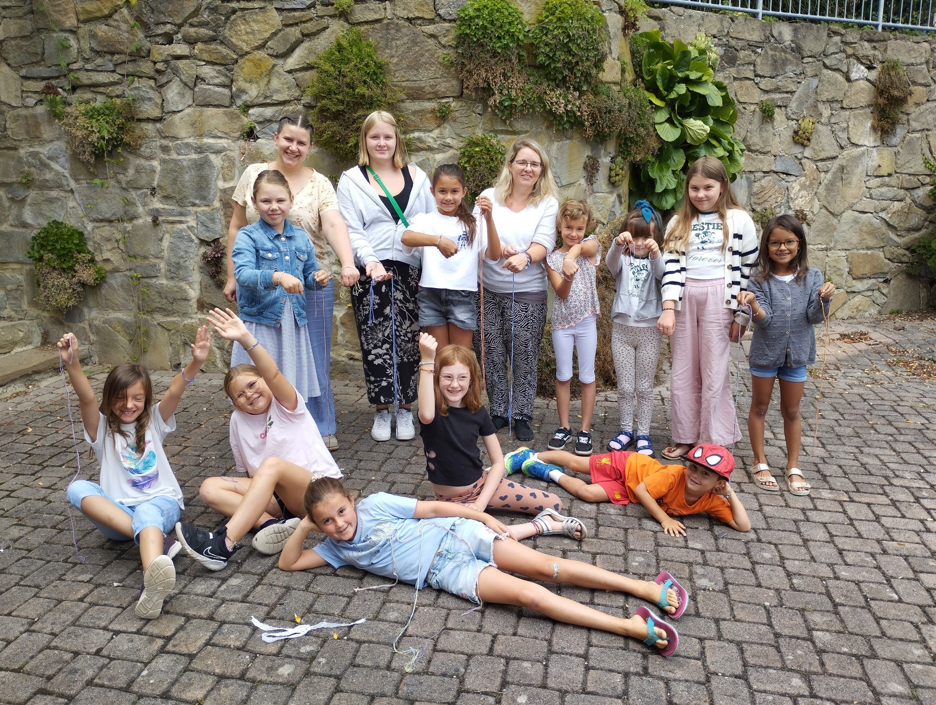 Kinder und Erwachsene sitzen und liegen auf Pflaster. Einige heben die Hände. Die Gruppe ist vielfältig gekleidet.