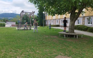 Kinder spielen Fußball auf einer Grünfläche mit einem Spielgerüst und einer Bank. Im Hintergrund ein Gebäude und Bäume.