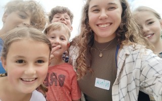 Mehrere Kinder und eine junge Frau lächeln vor der Kamera. Die Kinder tragen verschiedene Kleidungsstücke, darunter T-Shirts und Jeans. Die Frau trägt ein gemustertes Hemd über einem schwarzen Top und eine schwarze Umhängetasche.