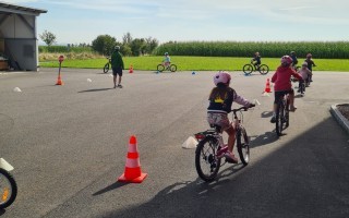 Kinder fahren Fahrrad auf einem asphaltierten Feldweg mit gelochterggeichen Maisfeldern im Hintergrund. Es sind gelbe Sicherheitsschilder und orangefarbene Cone zur Absicherung verteilt.