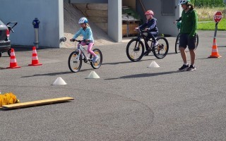 Kinder üben Fahrradfahren auf einer asphaltierten Fläche. Zwei Erwachsene und ein Kind fahren Fahrräder. Es stehen rote Warnsignale und eine Holzleiste auf dem Platz.