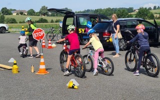 Kinder lernen Fahrradfahren auf einem Parkplatz mit Absperrpfosten und einem geöffneten Auto im Hintergrund.