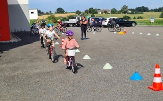Kinder lernen Fahrrad fahren auf einem Parkplatz mit leuchtenden Warnsignalen. Im Hintergrund Gebäude und Bäume.