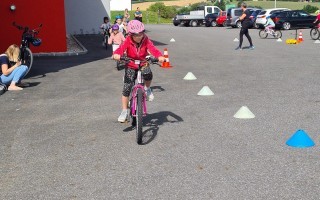 Kinder fahren Fahrrad auf einem Parkplatz mit blauen Hindernispirallen. Eine kleine Mädchen sitzt auf einem pinken Fahrrad und andere Kinder stehen am Rand.