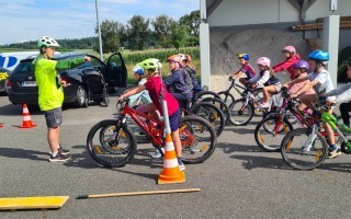 Kinder lernen Fahrrad fahren unter Aufsicht eines Trainers auf einem Feldweg mit einem dunklen Kleinwagen im Hintergrund.