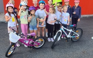 Mehrere Kinder fahren Fahrräder auf einer Straße. Einige tragen Helme. Der Hintergrund ist eine graue Wand mit Fenstern.