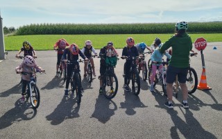 Kinder fahren Fahrrad auf einem asphaltierten Weg unter Aufsicht eines Erwachsenen. Im Hintergrund ein Maisfeld.