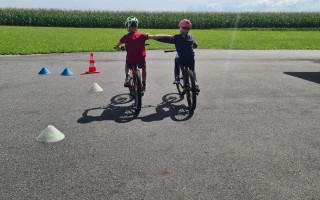 Zwei Kinder lernen Fahrradfahren auf einem asphaltierten Platz mit gelben Konischern. Ein Kind hilft dem anderen.